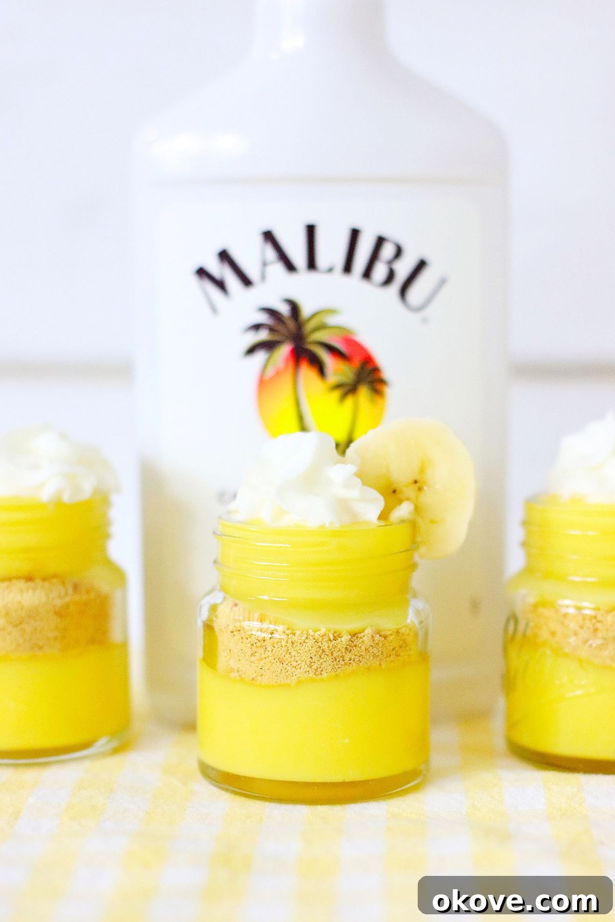 Sweet Banana Pudding Shooters 6 Beautifully layered banana pudding shots in mini jars, topped with a swirl of whipped cream and a fresh banana slice, with a bottle of Malibu rum visible in the background.