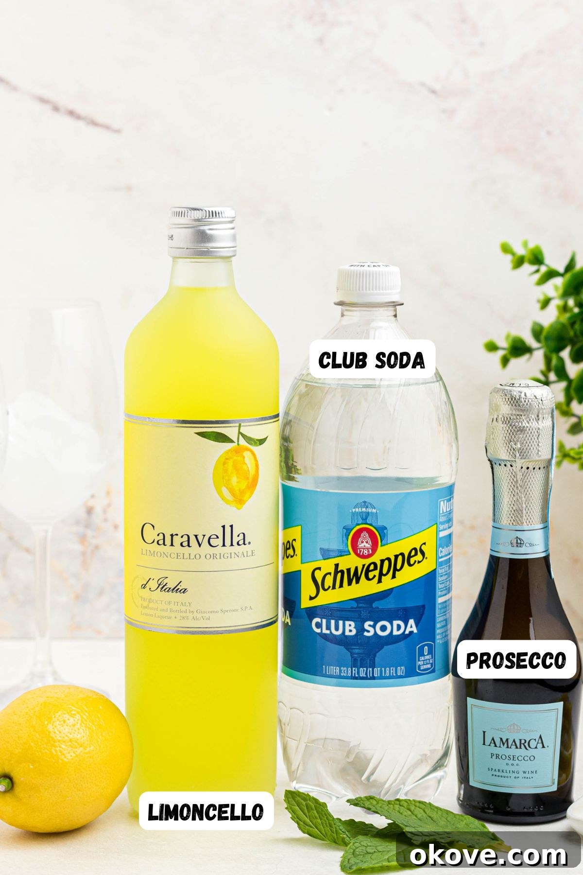 Limoncello Fizz 4 Ingredients for Limoncello Spritz laid out: bottles of limoncello, soda water, and prosecco, alongside fresh lemons and mint sprigs on a white marble table.