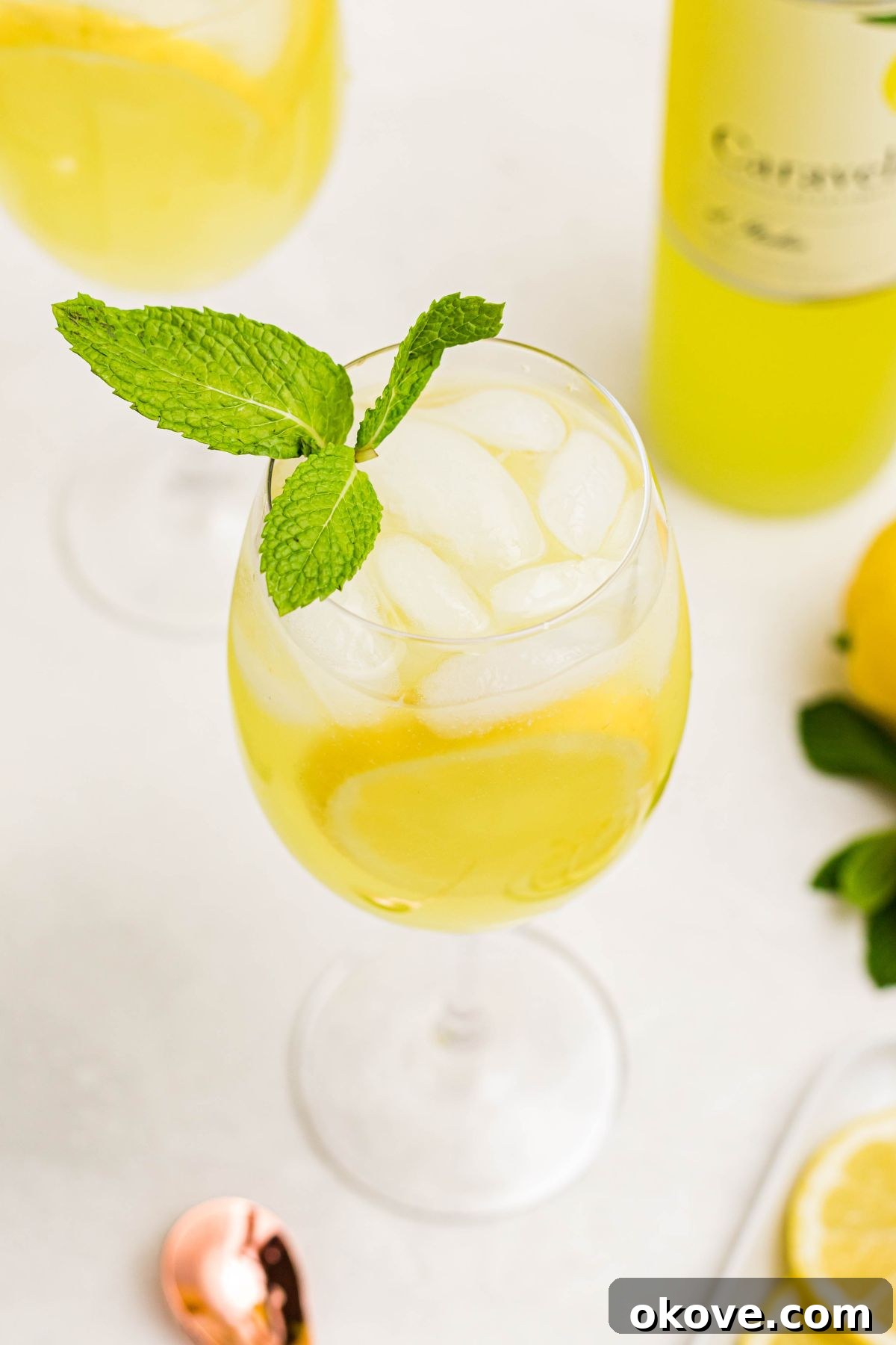 Limoncello Fizz 7 Image of a wine glass filled with a sparkling limoncello spritz, topped with a sprig of mint, resting on a white marble table.
