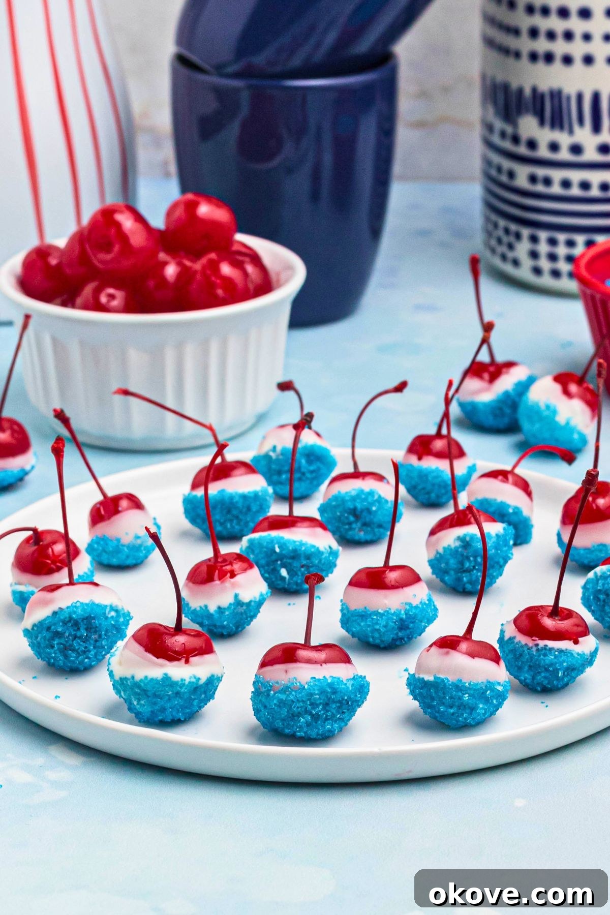 Bright red cherries coated in white chocolate and dipped in blue sanding sugar, on a white plate, with a bowl of cherries and blue coffee mugs in the background. 