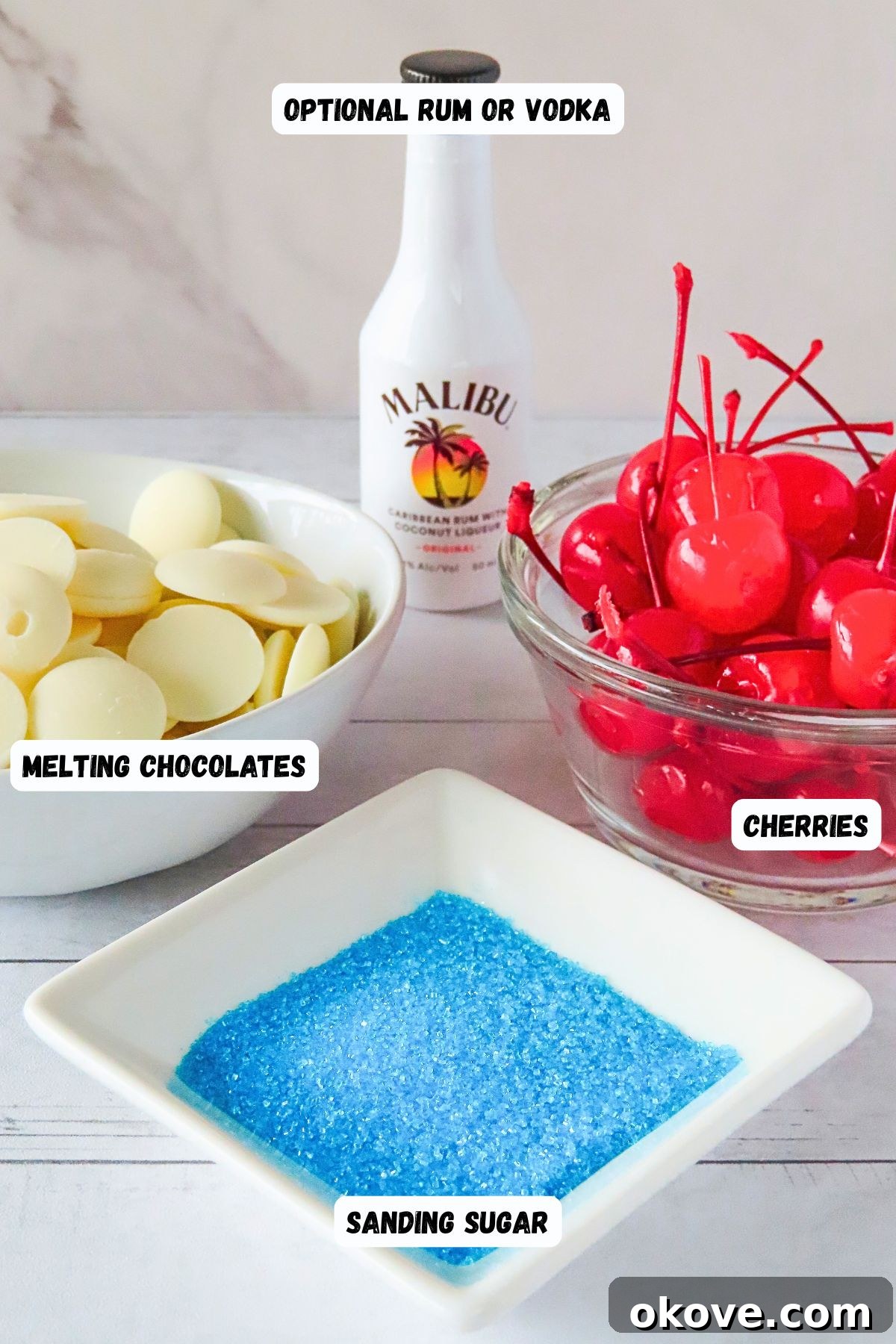 Cherries, white chocolate, sky blue sanding sugar, and optional small bottle of run, on a white wooden tabletop. 