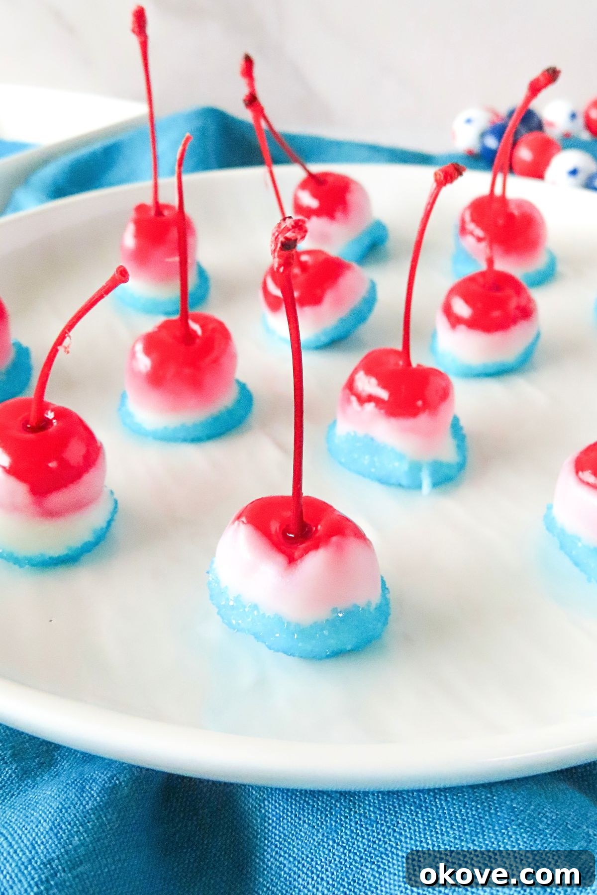 Red cherries, dipped in white chocolate, then dipped again in blue sanding sugar, on a plate waiting to dry. 