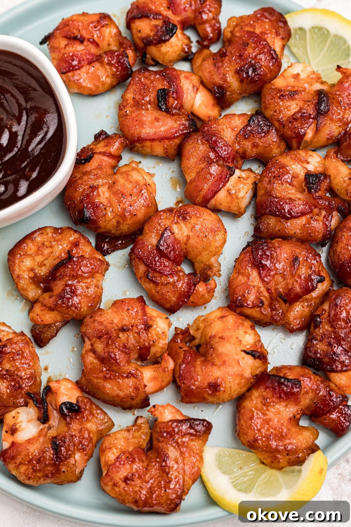 Crispy Bacon-Wrapped Shrimp Skewers with Garlic Butter Dip 2 Crispy bacon wrapped around shrimp, coated with BBQ sauce mixture, on a light green plate with lemon slices and serving tongs on the table.