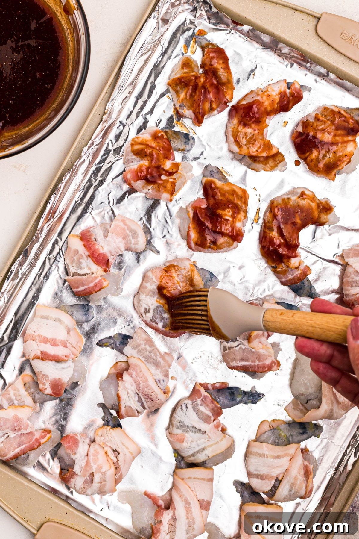Crispy Bacon-Wrapped Shrimp Skewers with Garlic Butter Dip 6 Shrimp wrapped in bacon, being brushed with BBQ sauce mixture, on a baking sheet lined with foil.