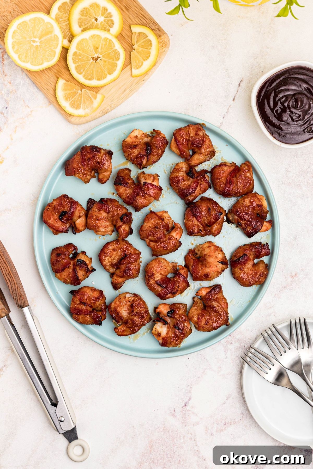 Crispy Bacon-Wrapped Shrimp Skewers with Garlic Butter Dip 7 Crispy bacon wrapped around shrimp, coated with BBQ sauce mixture, on a light green plate with lemon slices and serving tongs on the table.