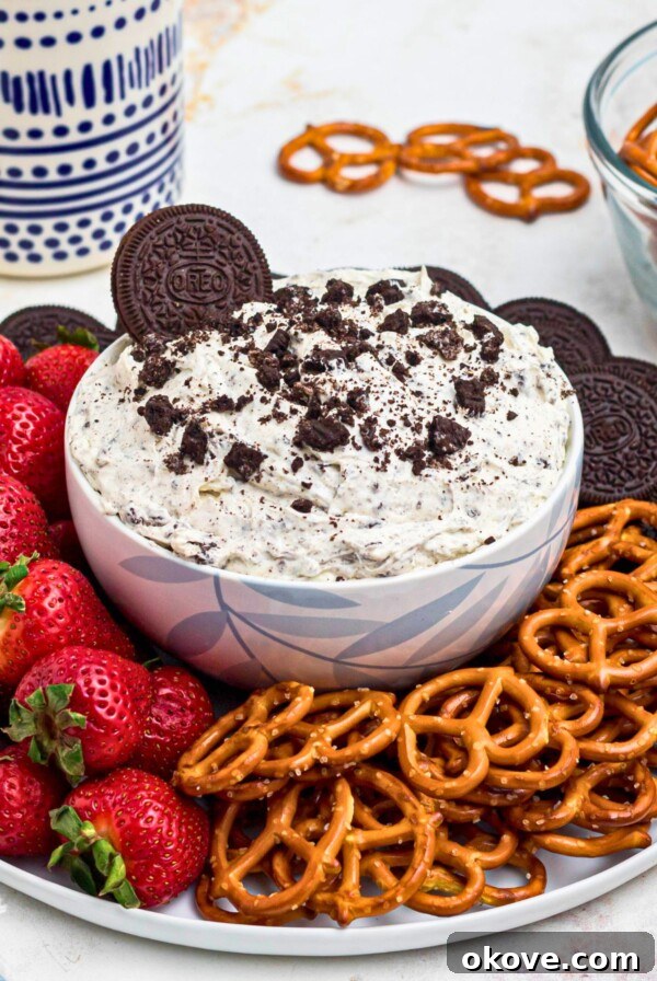 Strawberries, pretzels, and chocolate cookies, on a round white plate, around a cookies and cream Oreo dip, on a marble table.