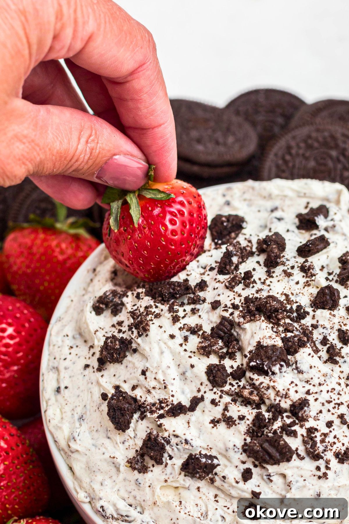 Creamy Oreo Dessert Dip for Parties and Snacks 8 A hand dipping a strawberry into Oreo dip, surrounded by more strawberries, Oreos, and pretzels.