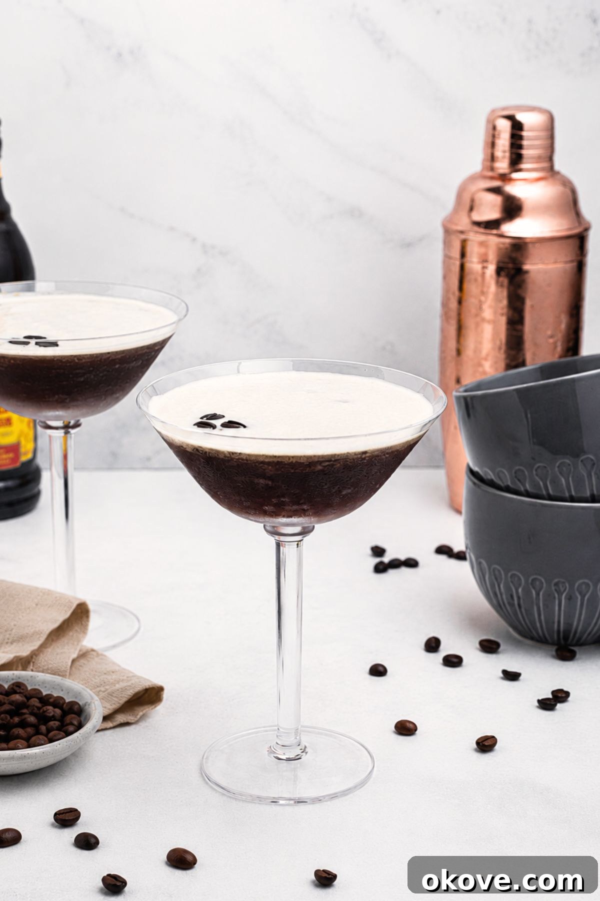 Espresso Martini Cocktail Recipe 2 Martini glass with coffee and vodka mixture, with a frothy foam on top, on a marble table with coffee beans scattered.