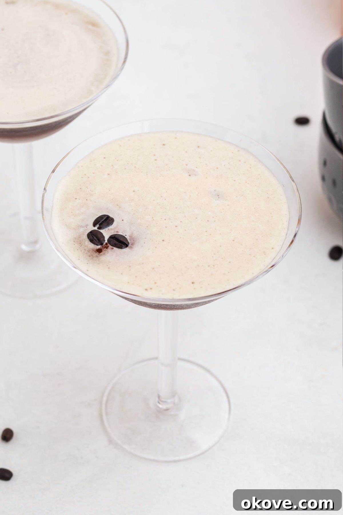Espresso Martini Cocktail Recipe 6 Froth on top of an espresso and vodka mixture, in a martini glass, with coffee beans spread around the table, and three coffee beans on top of the foam.