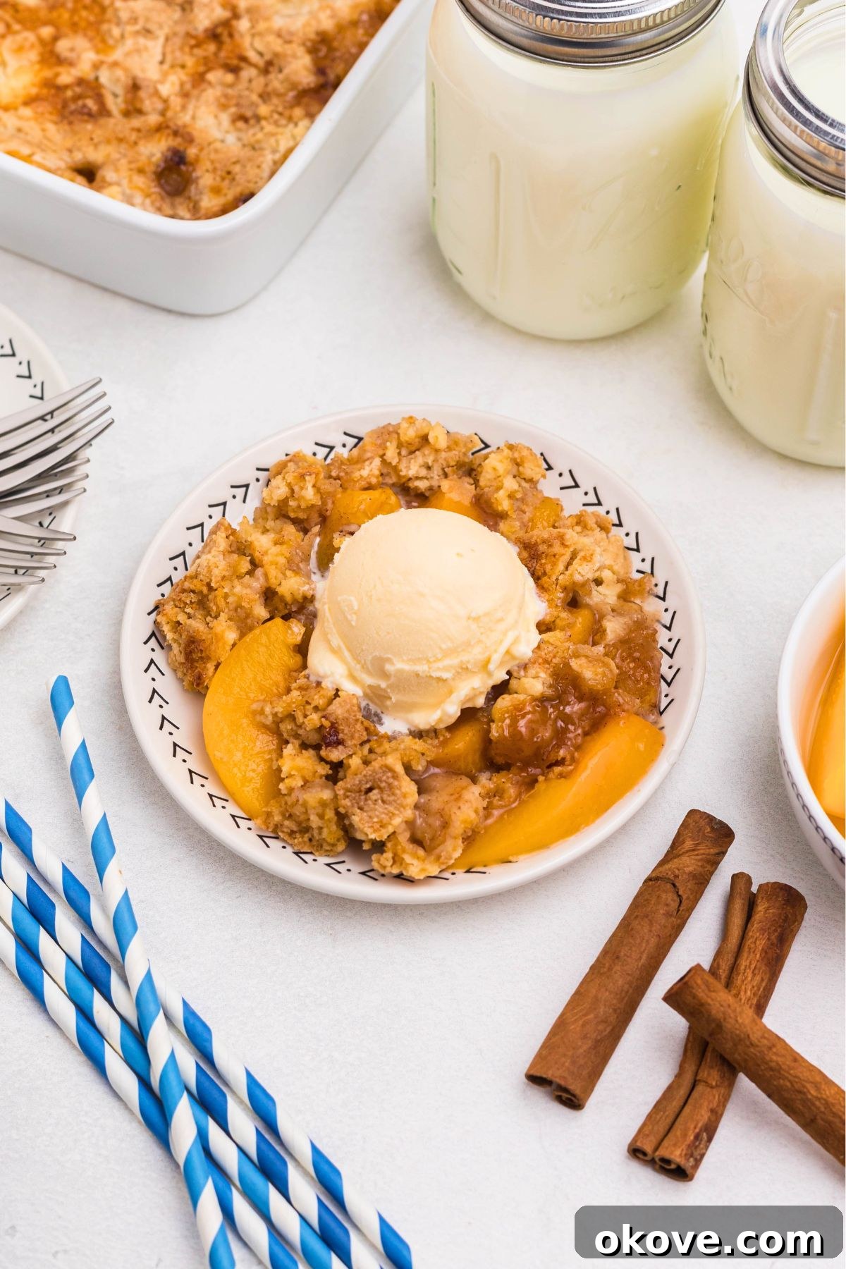 Irresistible Peach Dump Cake Recipe for Summer Desserts 8 Peach cobbler scooped out and served on a small plate, in front of the whole dish, with blue striped straws and cinnamon sticks on the table.