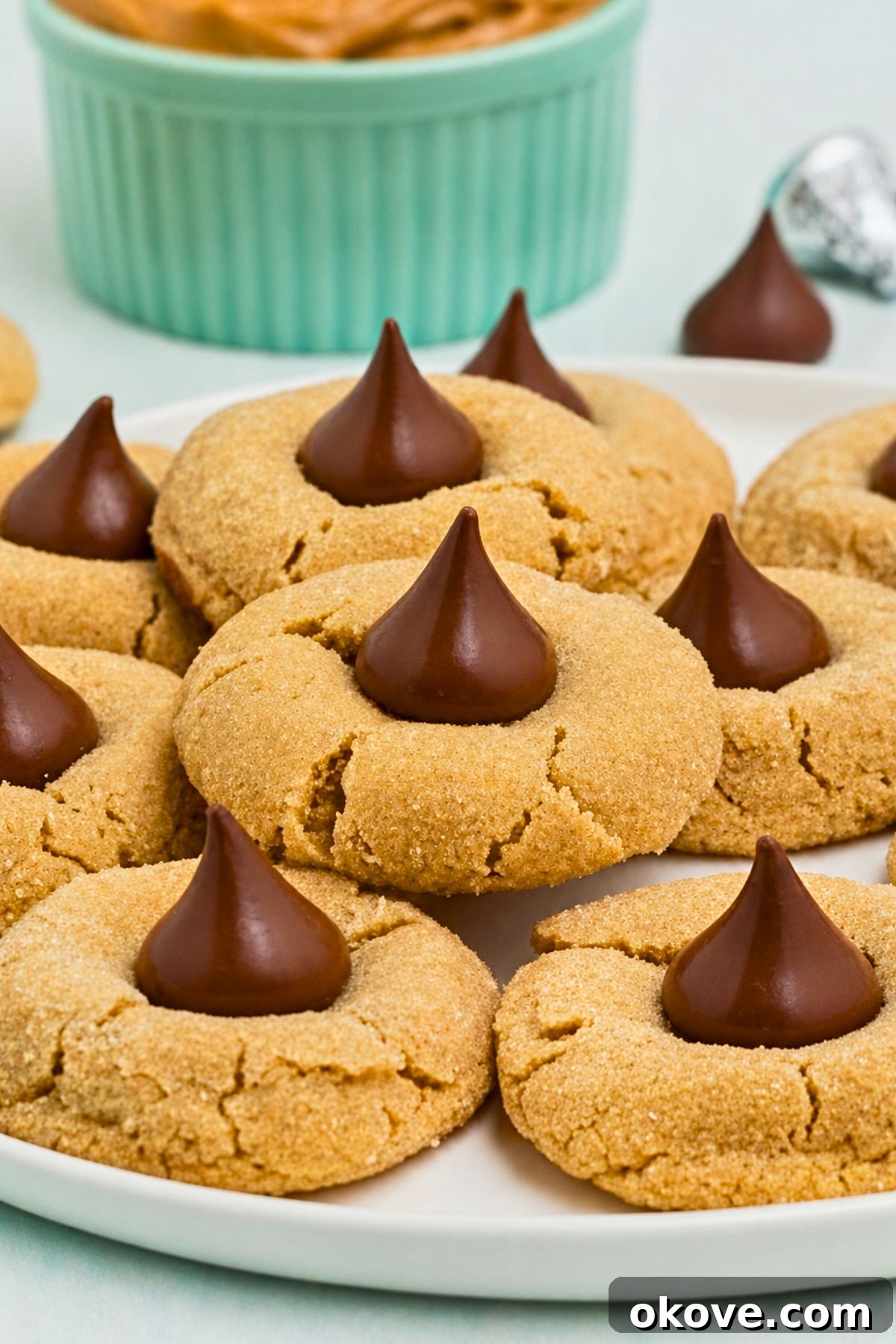 Classic Peanut Butter Blossom Cookies with Chocolate Kiss 2 Peanut Butter cookies, topped with a chocolate kiss, stacked on a white plate, with a bowl of peanut butter on the table.