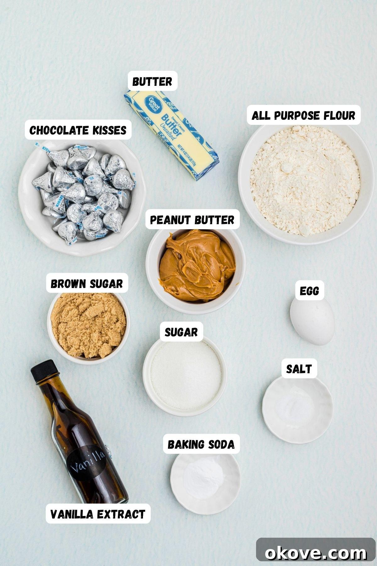 Classic Peanut Butter Blossom Cookies with Chocolate Kiss 3 Creamy peanut butter, Unsalted butter, Granulated sugar, Light Brown sugar, Egg, Vanilla extract, All-purpose flour, Baking soda, Salt, and Hershey's, Kisses in small bowls, on a light blue table, with all ingredients labeled.