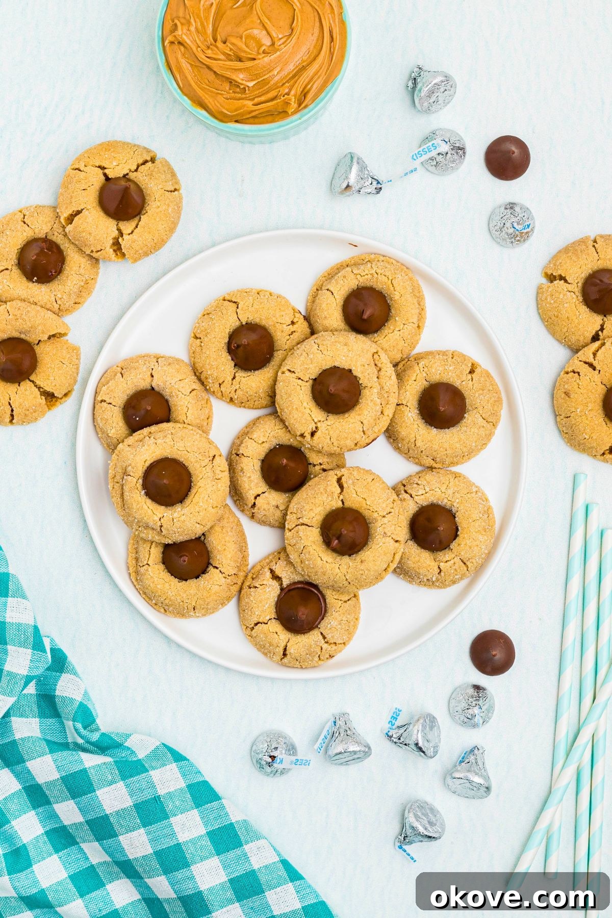 Classic Peanut Butter Blossom Cookies with Chocolate Kiss 6 Peanut Butter cookies, topped with a chocolate kiss, stacked on a white plate, with a bowl of peanut butter on the table, next to a teal checked linen.