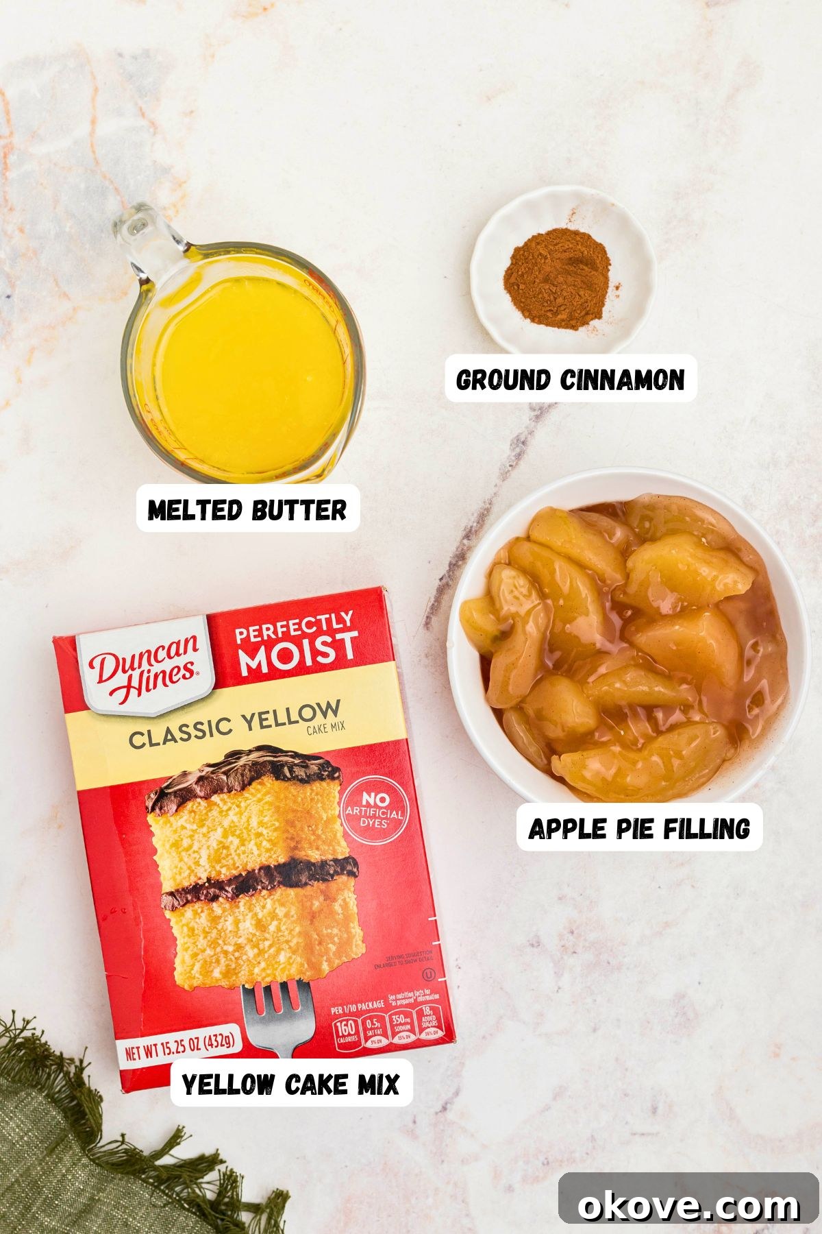 Box cake mix, melted butter, cinnamon, and apple pie filling, on a marble table, labeled, with a green linen on the table.