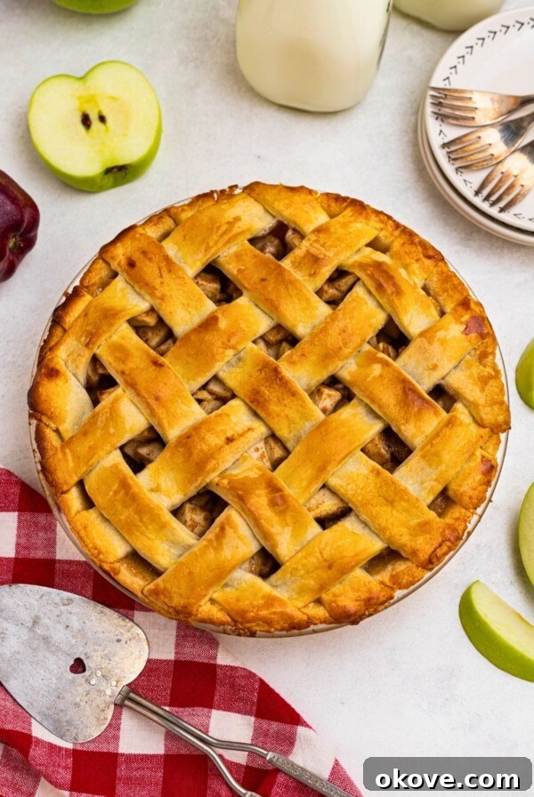 Golden crust, baked apple pie, on a white marble table, with slices of green apple, and a small plate with forks, next the whole pie.