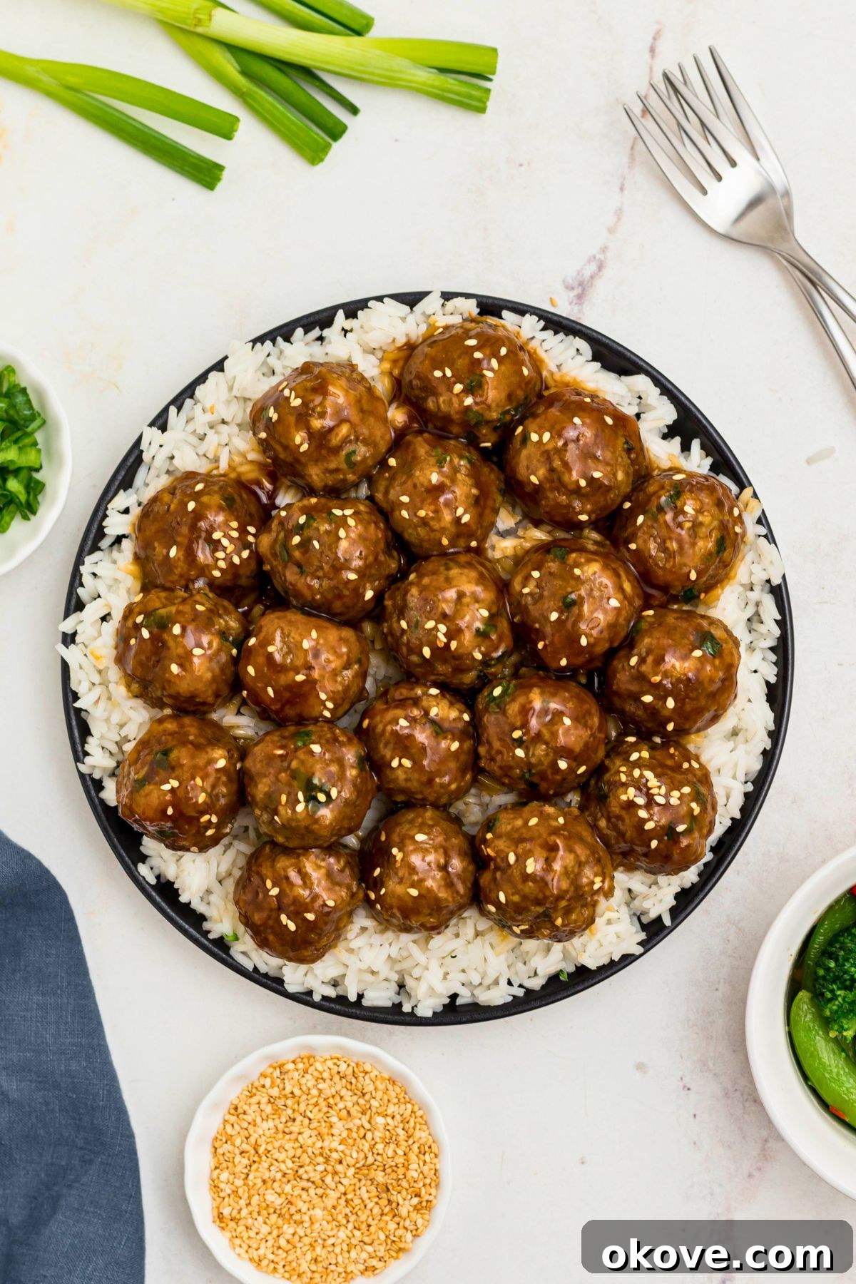 Black plate topped with white rice and juicy meatballs surrounded by chopped onions and sesame seeds on a marble table.