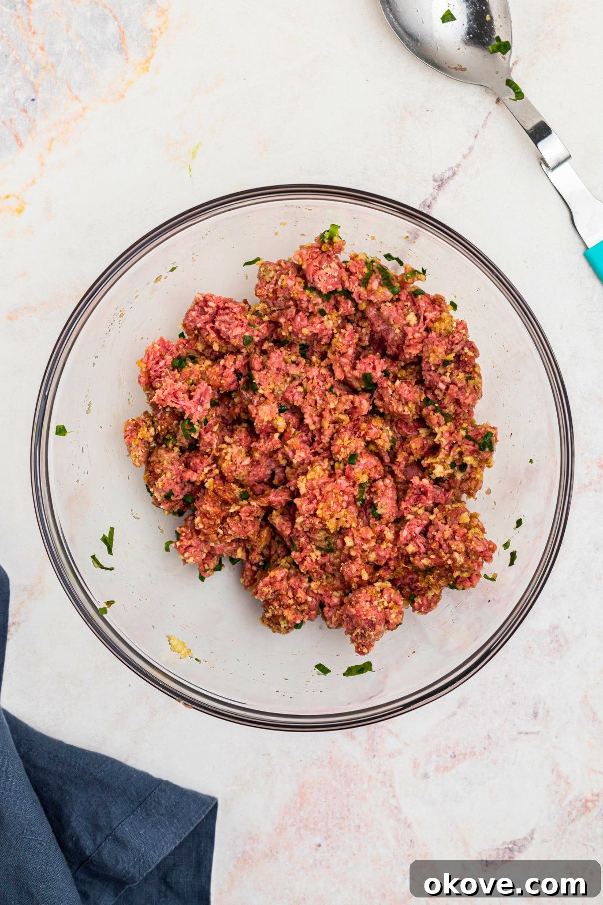 Ground beef mixed with other ingredients, in a large clear glass bowl, with a blue linen on a marble table.
