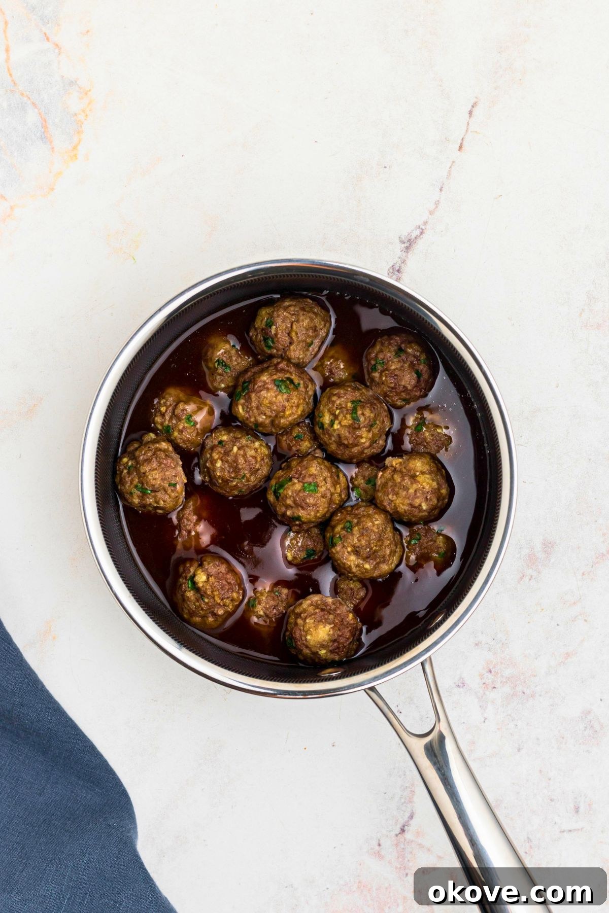 Saucepan filled with teriyaki sauce and cooked meatballs, on a white marble table with a blue linen on the table.