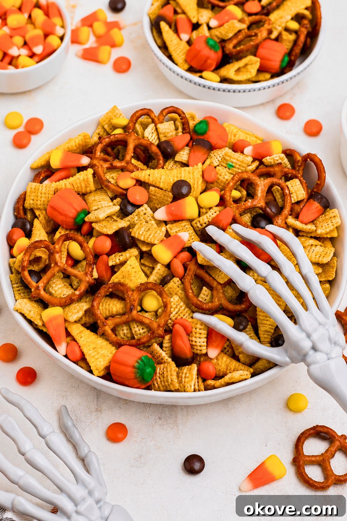 Spooky Halloween Chex Mix with Candy Corn and M&Ms 2 Large white bowl filled with Chex mix, pretzels, candy corn, and candies, with some scattered around the table and a scooper skeleton hand on top of the bowl.