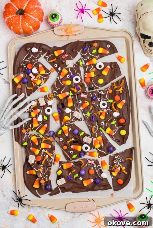 Spooky Halloween Chex Mix with Candy Corn and M&Ms 11 Halloween bark, broken into triangle pieces of chocolate, topped with candies, sprinkles, and candy eyes, on a baking sheet, with plastic spiders scattered around the table.