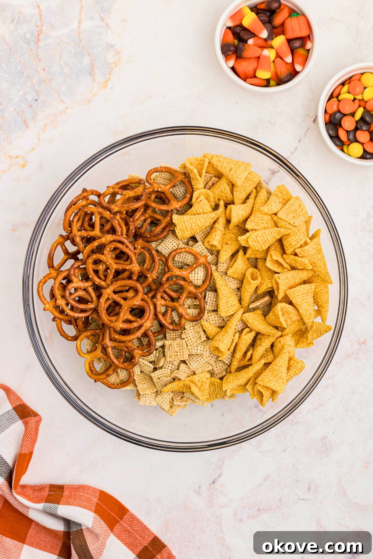 Spooky Halloween Chex Mix with Candy Corn and M&Ms 4 Cereal, pretzel, and bugles, in a large clear mixing bowl, with candies in small white bowls on the table.