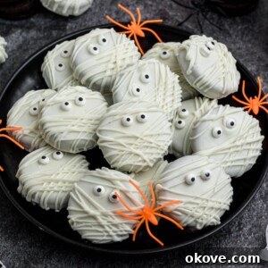 Oreo cookies, dipped and drizzled in white chocolate, with candy eyes, on a black plate, with orange spiders as decoration, on a black marble table.