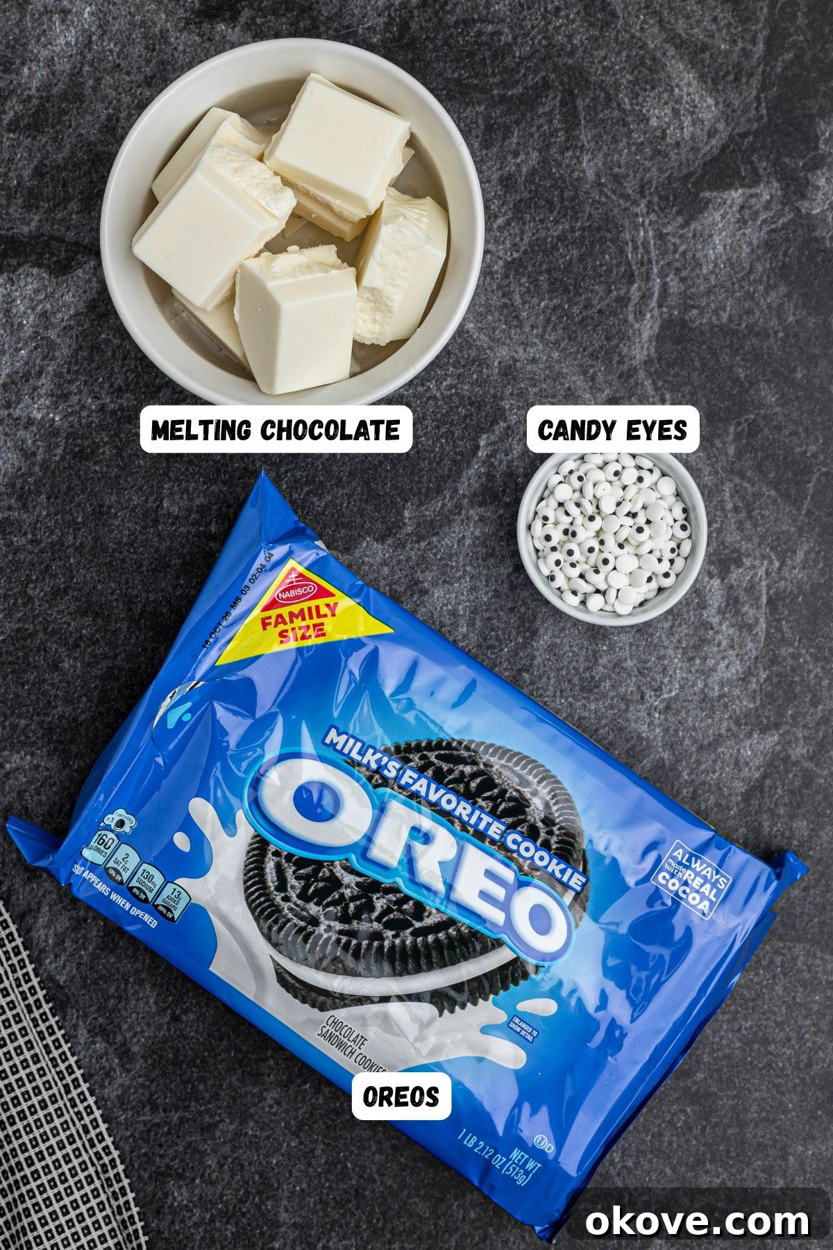 Oreo Mummy Cookies ingredients of Oreos, white chocolate, and candy eyes, on a black marble table, labeled. 