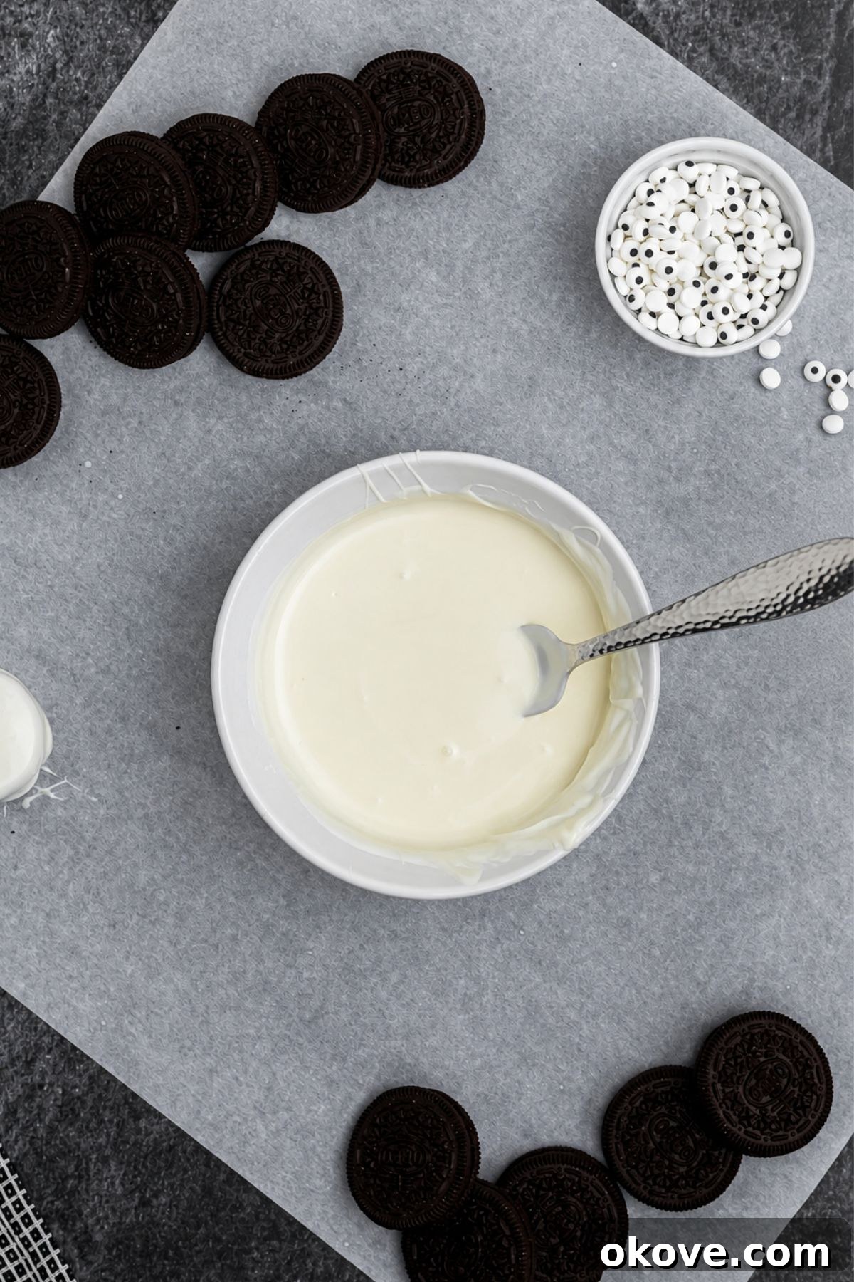 White chocolate melted in a small, shallow bowl, with Oreo cookies on the table, on parchment paper. 