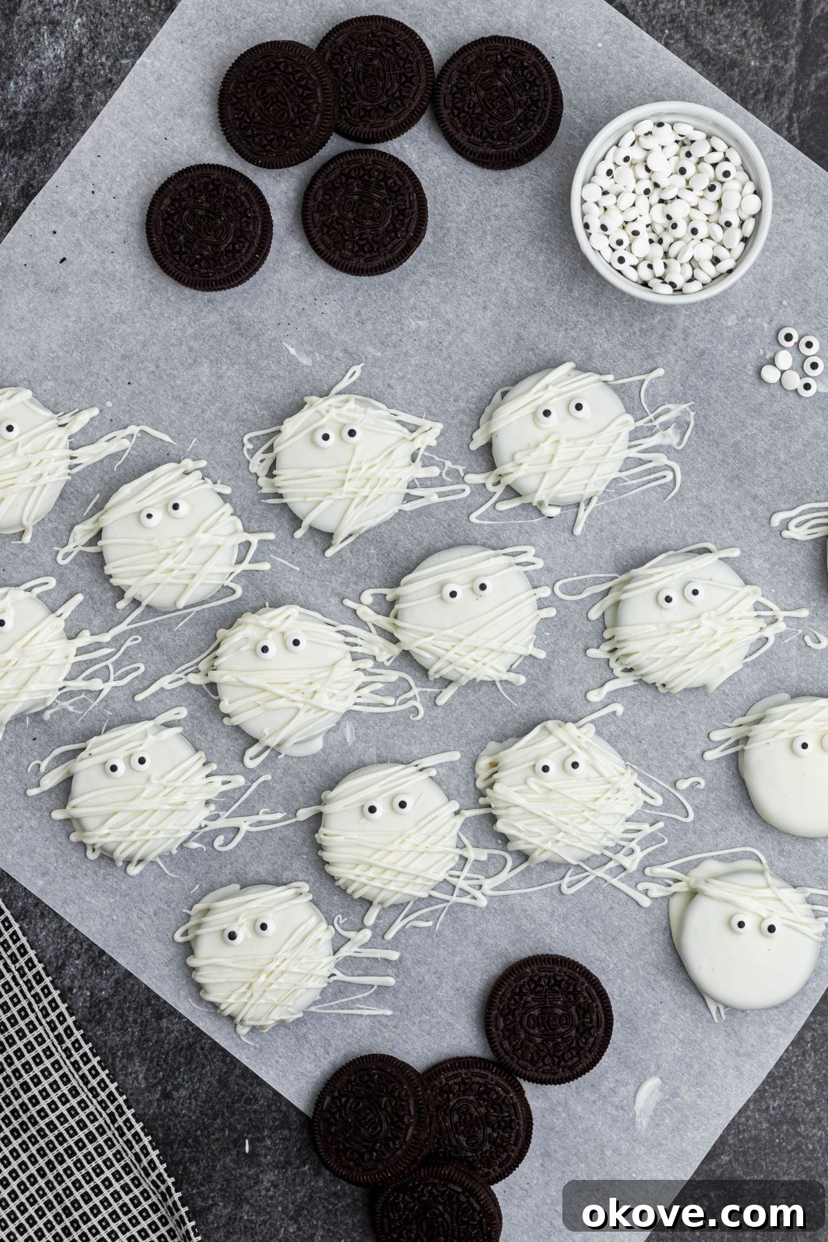 Oreos dipped in white chocolate, drying on parchment paper, with Oreos scattered on the table, with candy eyes added. 