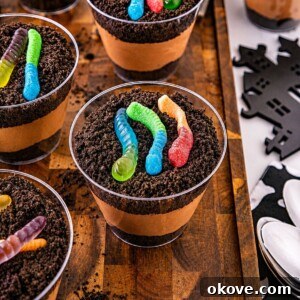 Small individual cups filled with crushed cookies, whipped cream, and chocolate pudding, topped with gummy worms, on a wooden cutting board.