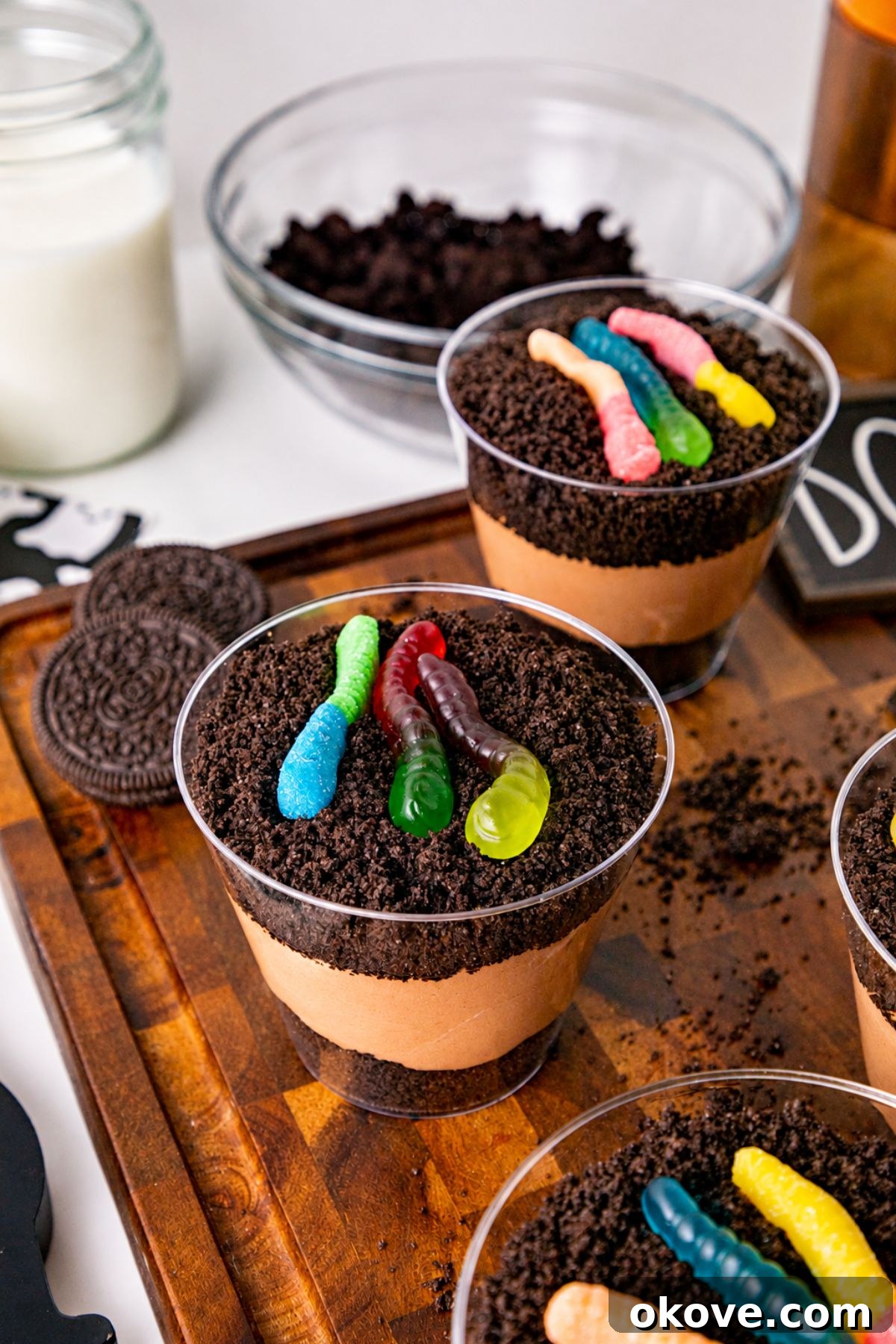 Small individual cups filled with crushed cookies, whipped cream, and chocolate pudding, topped with gummy worms, on a wooden cutting board.