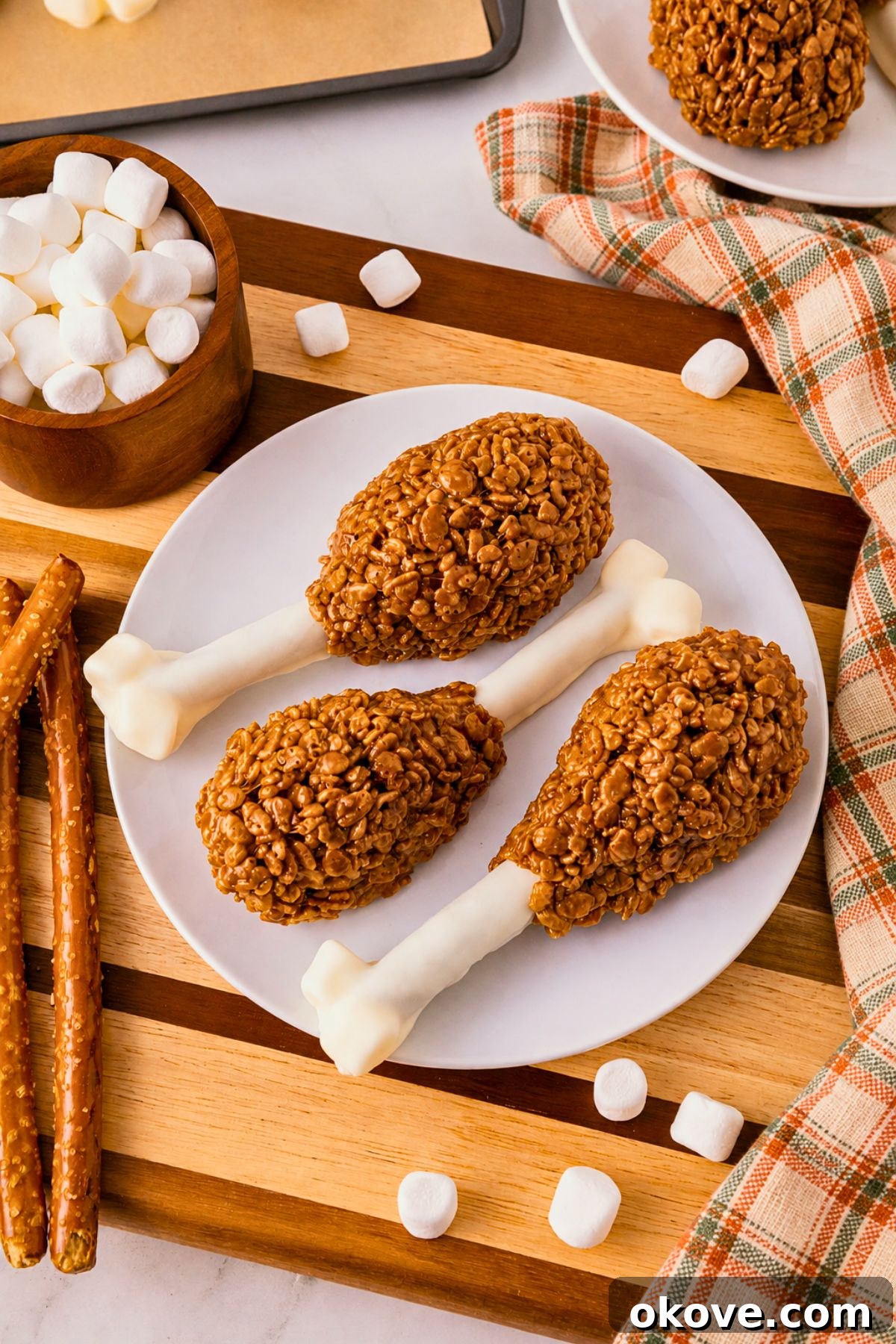 Cocoa krispie treats shaped around white chocolate dipped pretzel rods and marshmallow tips to look like turkey drumsticks. On a white plate, set on wooden cutting board, with marshmallows scattered around.
