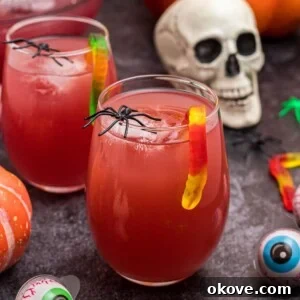 Glass of Halloween Punch in with gummy worms hanging over the side of the glass, in front of plastic skull and spiders.