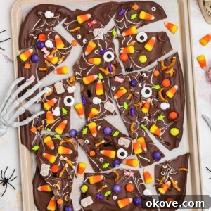 Halloween bark, broken into triangle pieces of chocolate, topped with candies, sprinkles, and candy eyes, on a baking sheet, with plastic spiders scattered around the table.