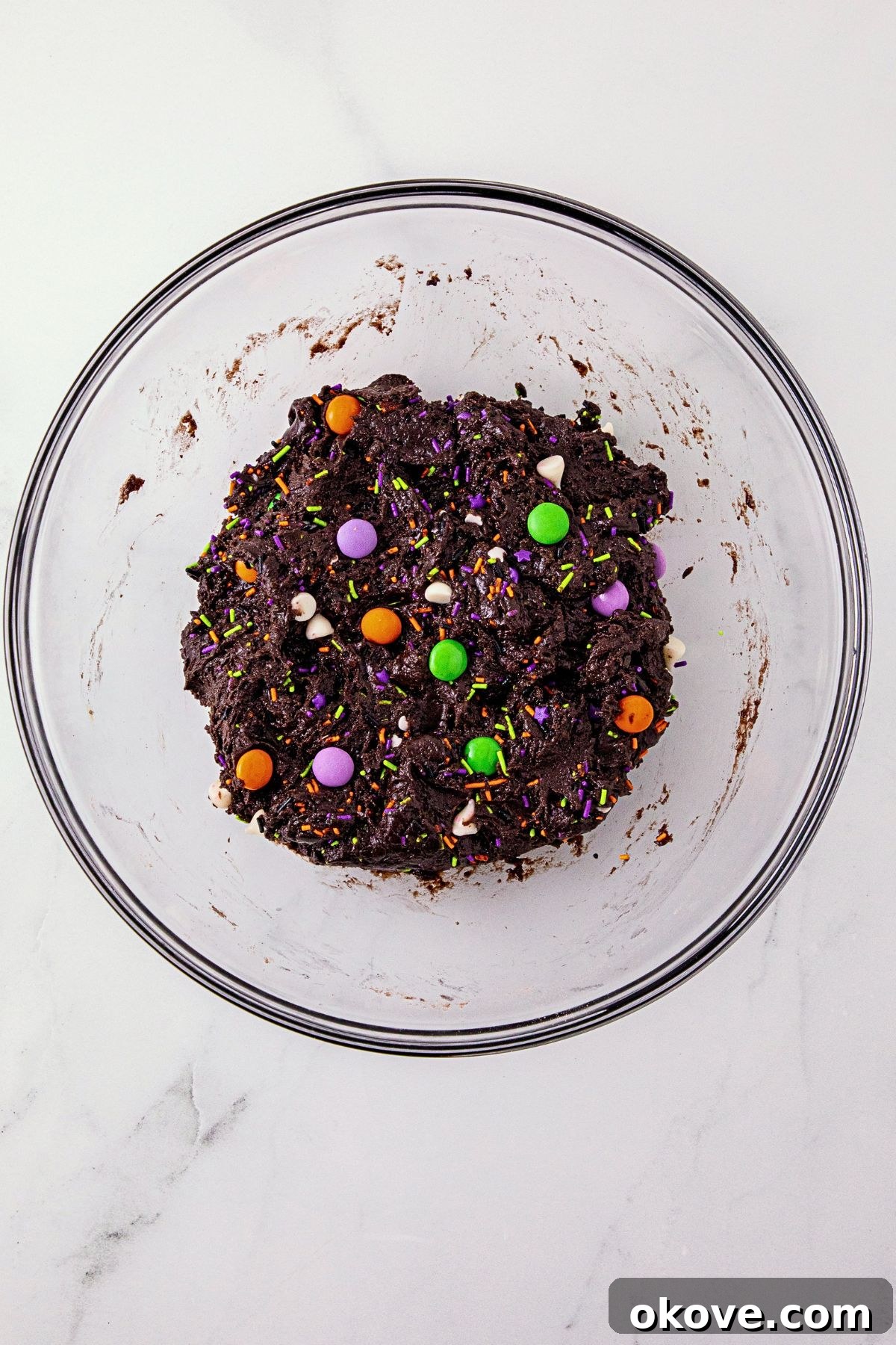 Chocolate cake mix cookie batter, in a large clear glass bowl, with Halloween sprinkles and candies mixed in. 