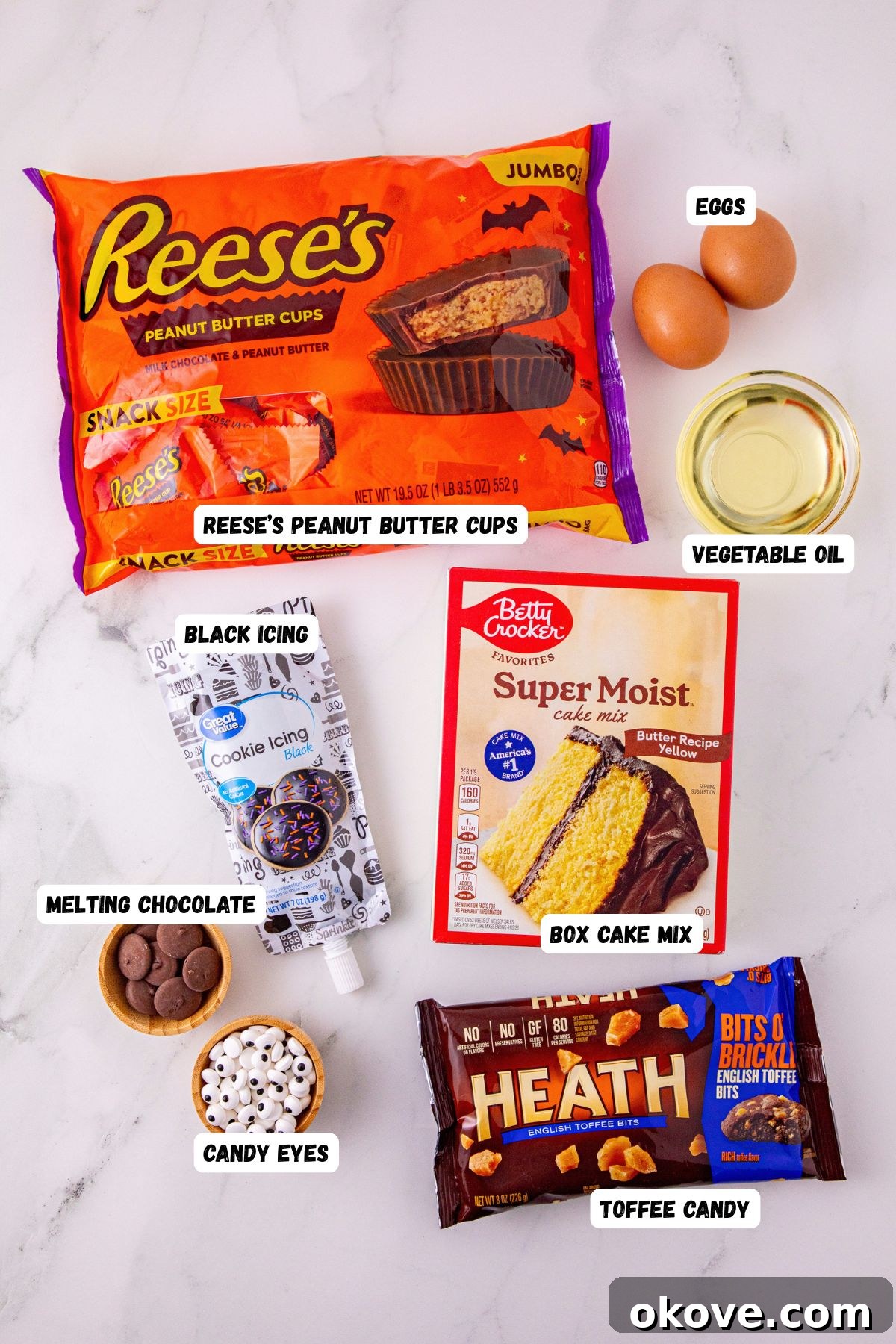Ingredients like cake mix, eggs oil, black icing, and candy eyes, on a marble table, labeled. 