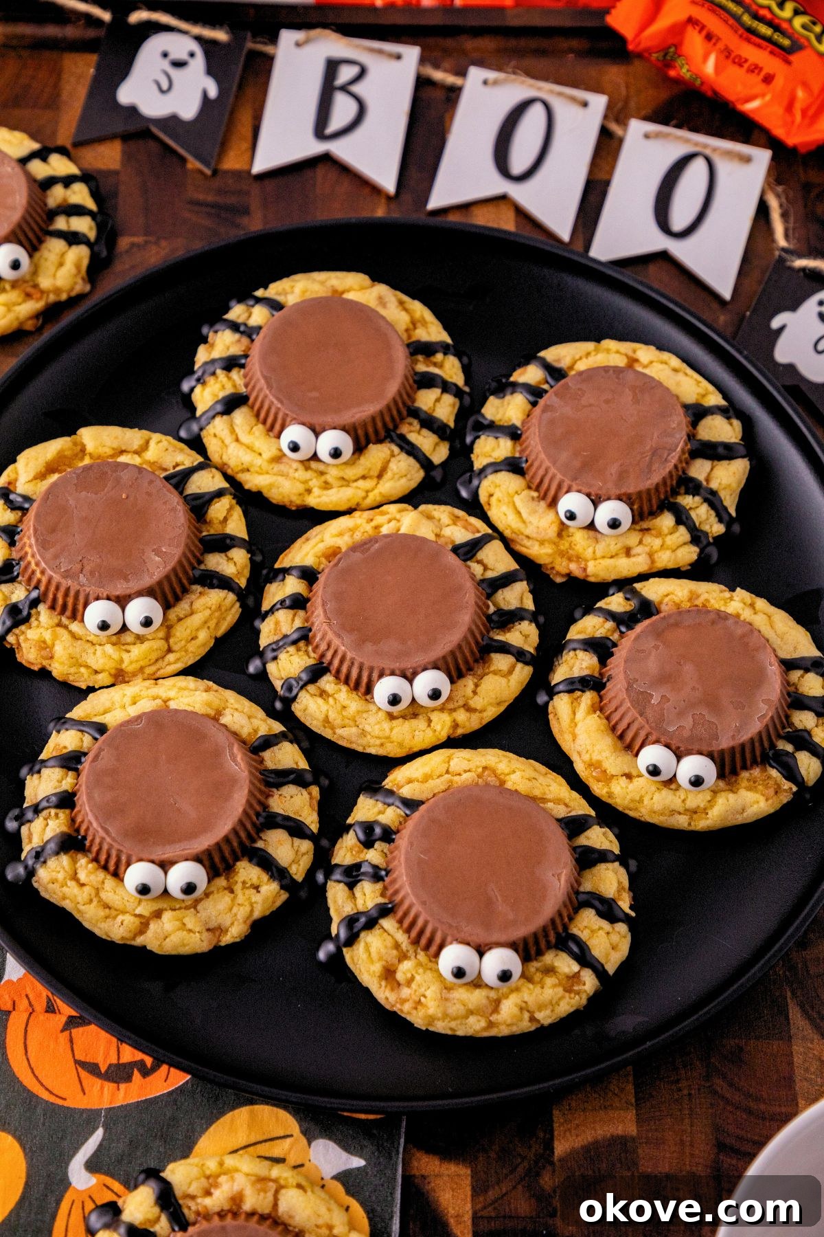 Black plate with spider cookies stacked around the plate, on a table with Halloween decorations scattered around the plate. 