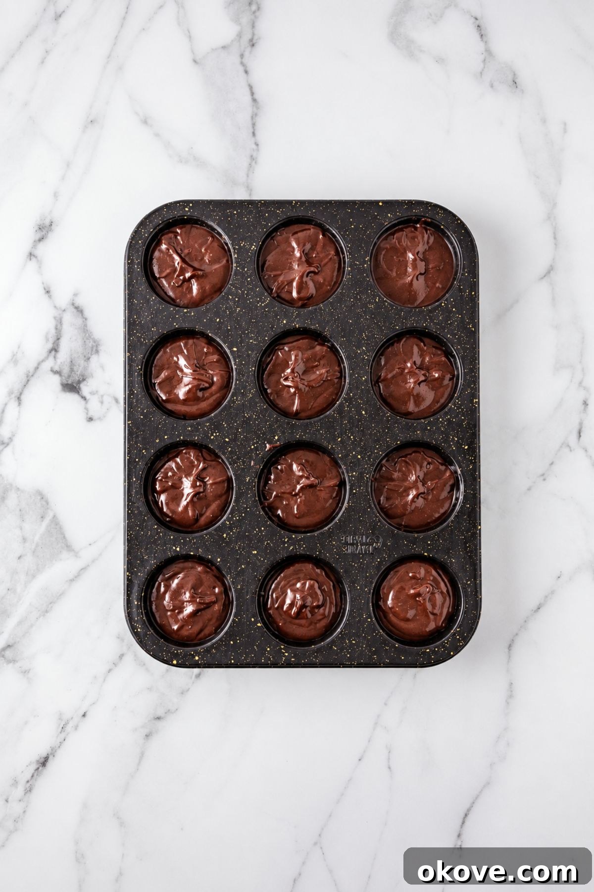 Muffin tin filled with brownie batter before baking, on a marble table. 