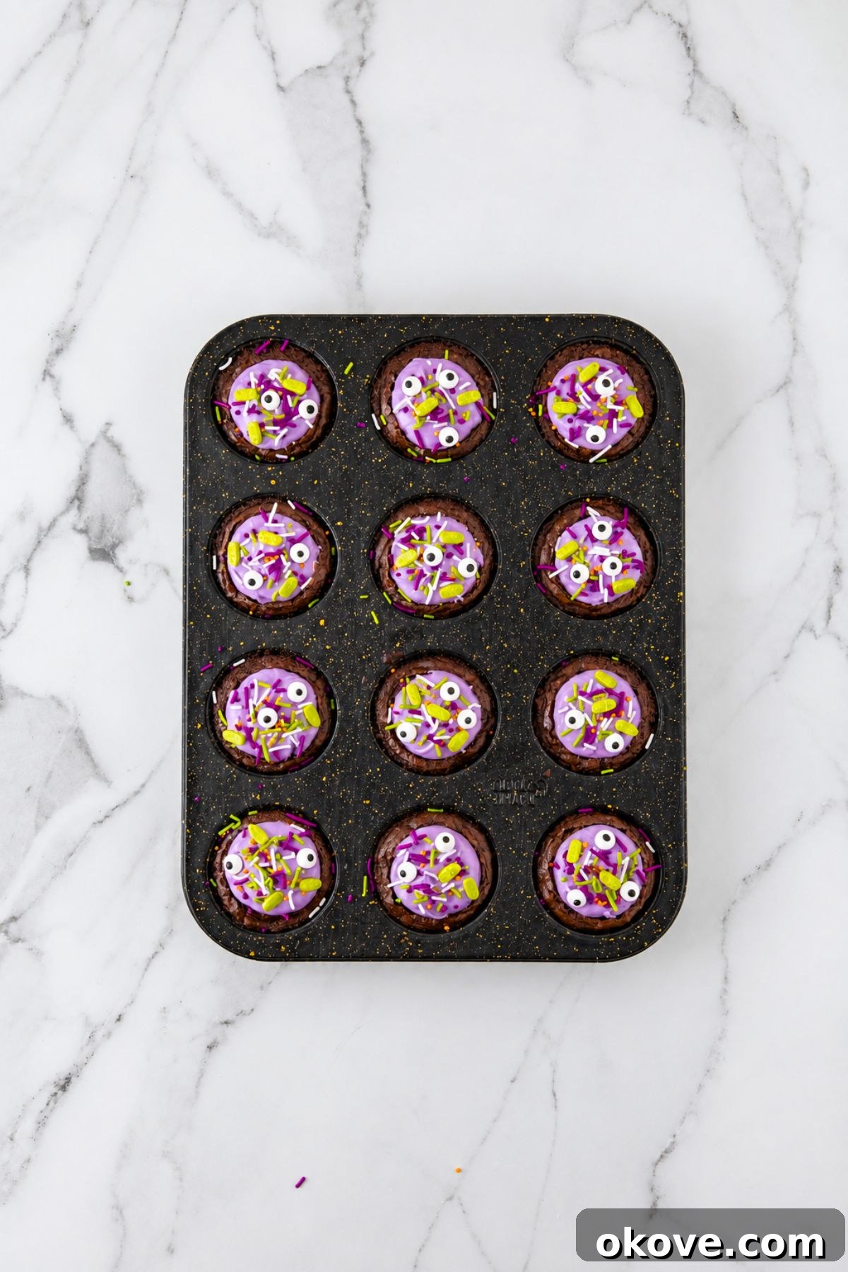 Brownie bites topped with purple frosting and sprinkles, in a muffin tin, on a marble table. 
